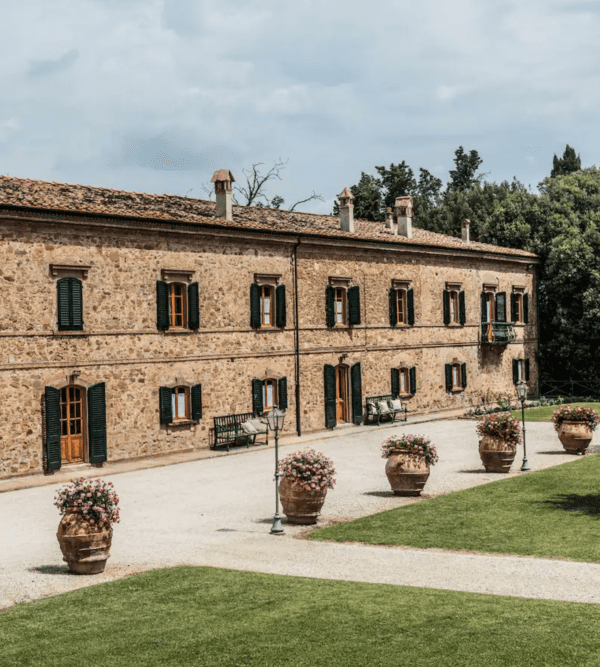 Historic stone building with green shutters, lined with large flower pots on a gravel pathway, bordered by lush greenery.