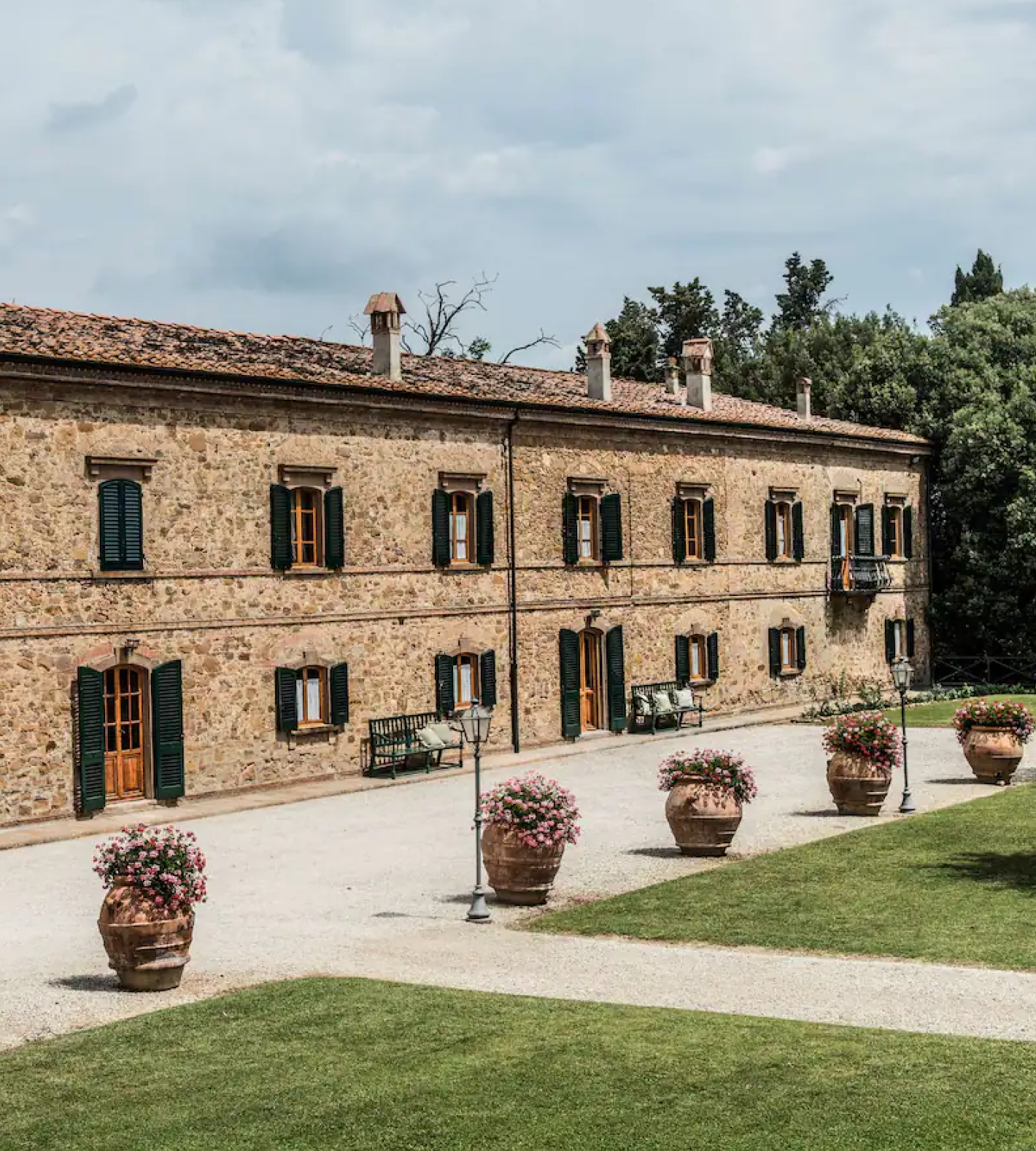 Historic stone building with green shutters, lined with large flower pots on a gravel pathway, bordered by lush greenery.