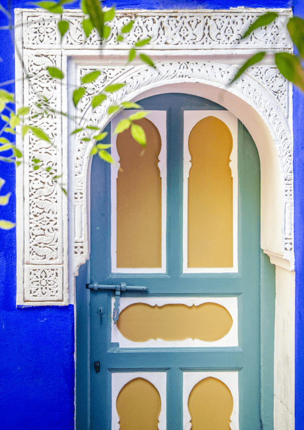 Colorful Moroccan door with ornate arch and vibrant blue wall, framed by green leaves.