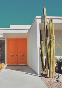 Mid-century modern house with vibrant orange doors and tall cacti in desert landscape under clear blue sky.