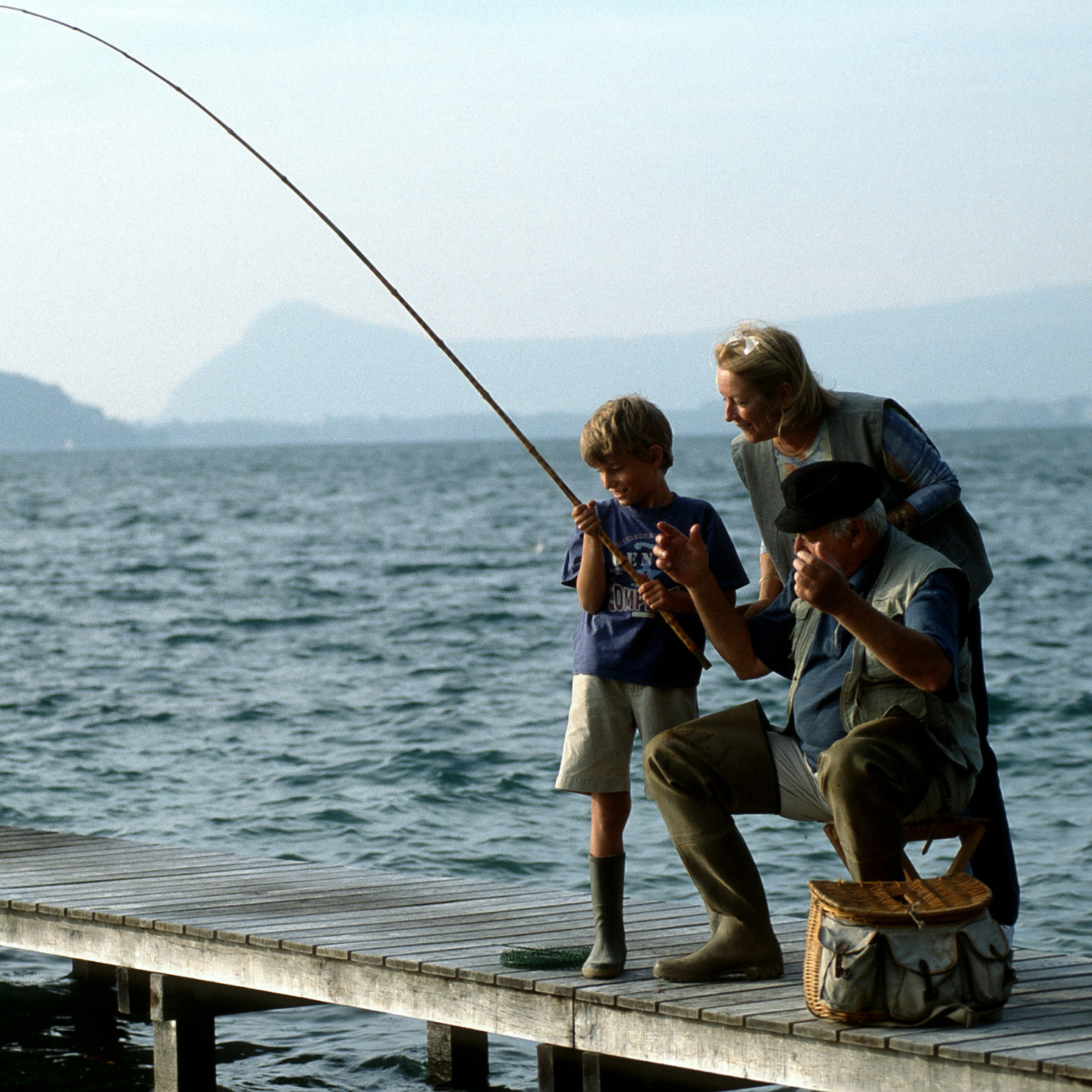 Family fishing on a dock: child reeling in a fish with guidance from elder and adult; scenic mountain lake backdrop.