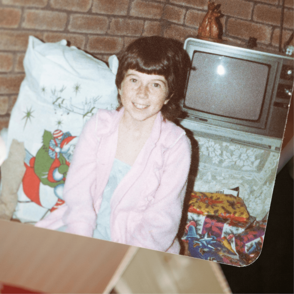 Smiling girl in pink robe sitting by vintage TV with gifts and festive background.