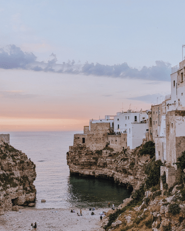 Coastal cliffs and historic buildings at sunset, with a serene beach below, in Polignano a Mare, Italy.