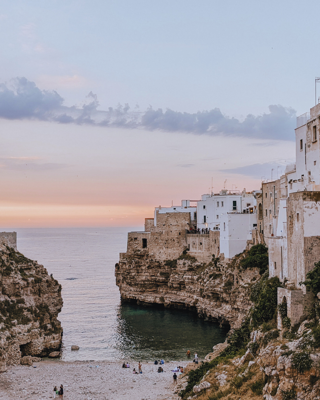 Coastal cliffs and historic buildings at sunset, with a serene beach below, in Polignano a Mare, Italy.